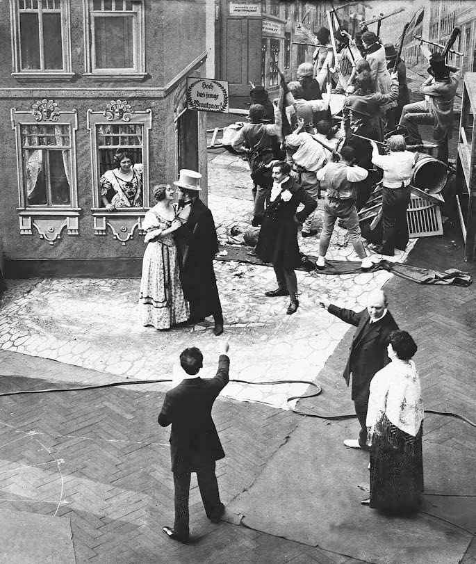 Open-air film set seen from above: a couple embrace outside a painted house facade while a crowd carrying rifles surges up the street behind them, and the crew watches from the foreground
