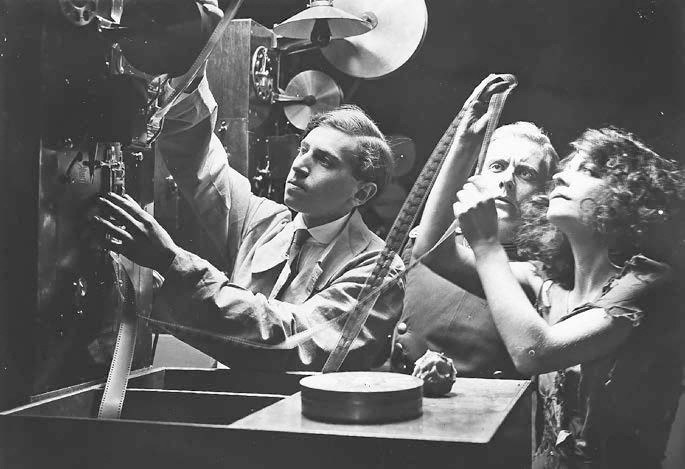 Film still: a young man in shirtsleeves feeds a long loop of film into a projector while two women lean in over his shoulder, watching intently