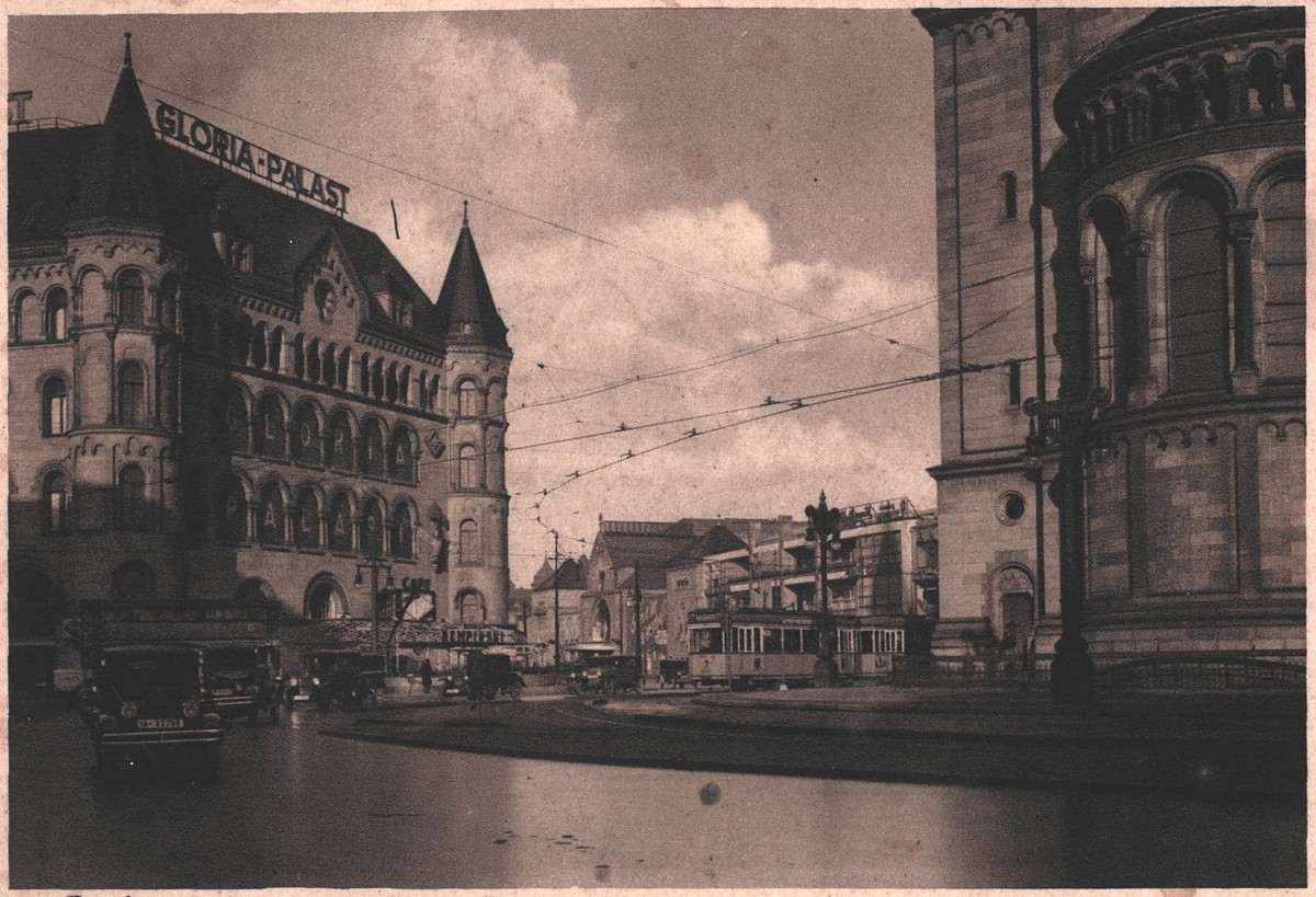 Photograph of a street corner with the turreted facade of the Gloria-Palast at left, its name in large letters along the roofline, overhead tram wires crossing the sky, and a tram and cars on wet cobbles