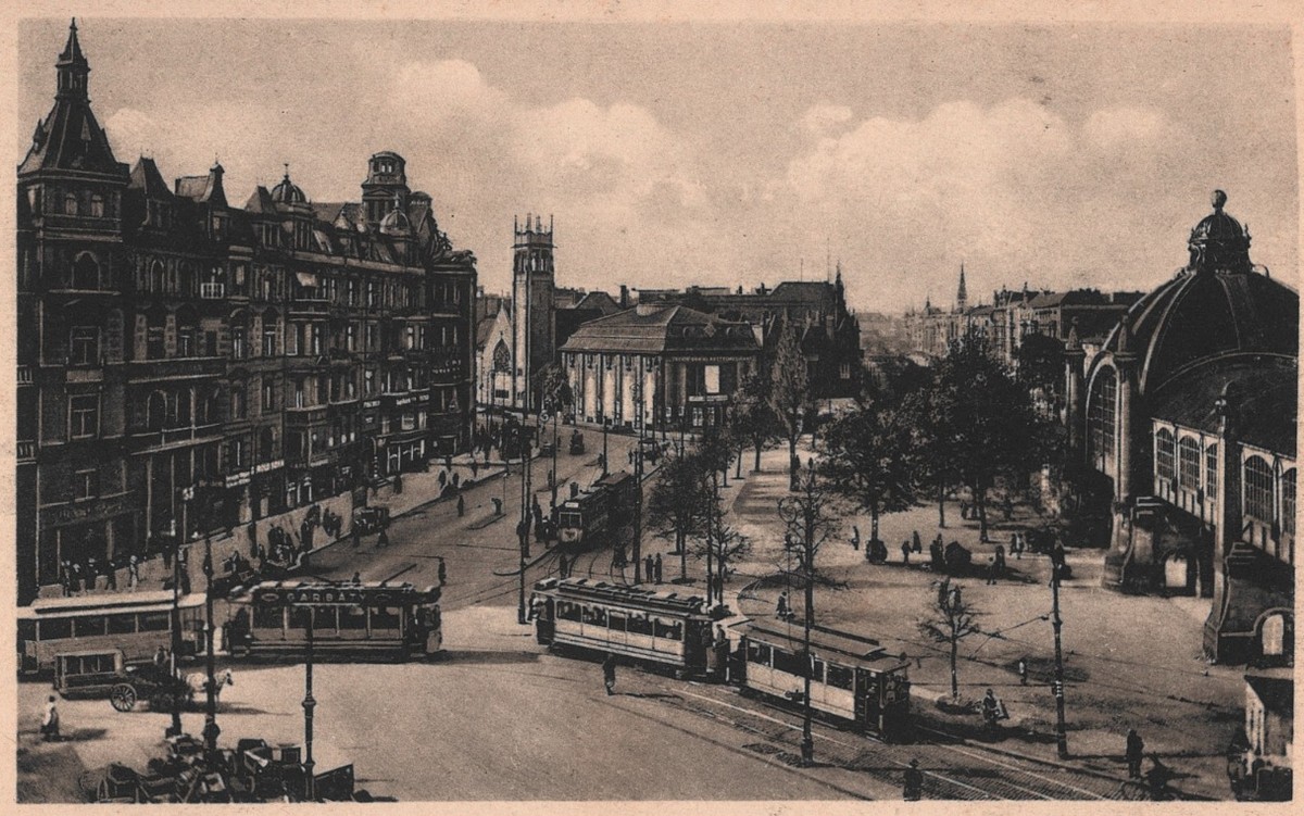 Photographic postcard of a wide square laid with tram tracks: apartment blocks along the left, a domed cinema building at right, and several trams and a horse cart crossing the open space