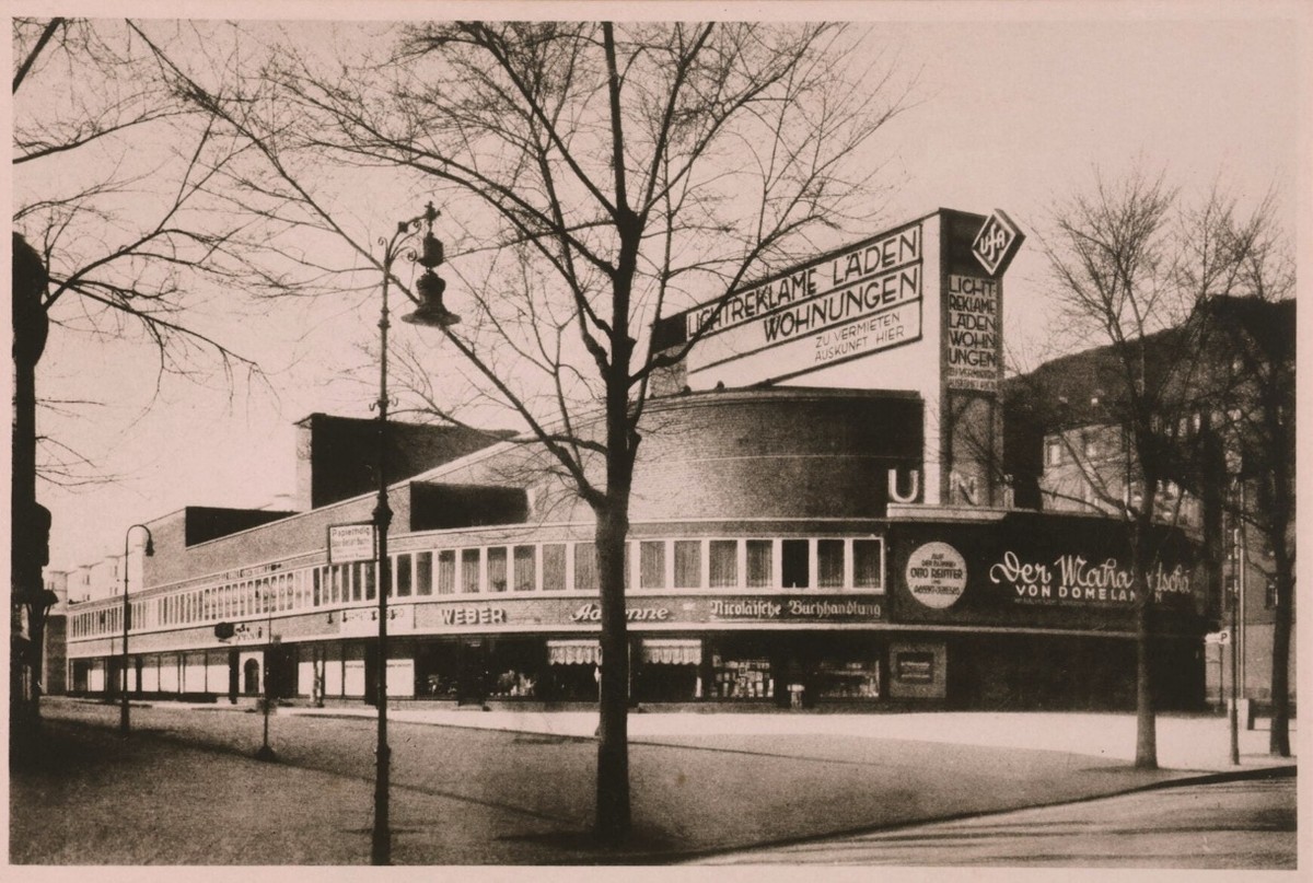 Photograph of a modernist cinema on a corner: a windowless curved drum beside a long glazed shopfront, an Ufa lozenge on the roof sign, a painted film poster on the side wall, and bare trees in front