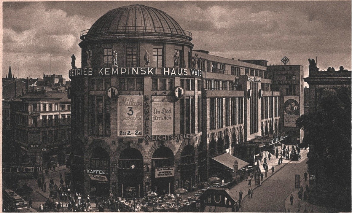 Photograph of a large domed building lettered "Betrieb Kempinski Haus Vaterland" along its roofline, hung with painted film posters and a Lichtspiele sign, with crowds and awnings at street level