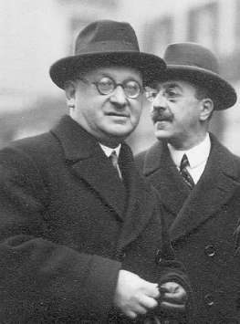 Two men in overcoats and hats outdoors, the nearer one in round glasses, the other with a moustache standing just behind him