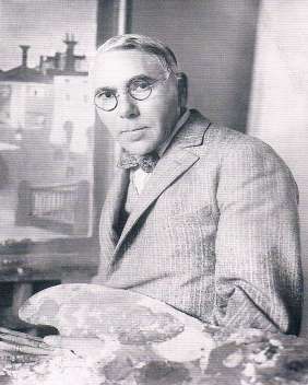 A man in round glasses, a light checked jacket and bow tie, seated in a studio holding a painter's palette with a canvas behind him