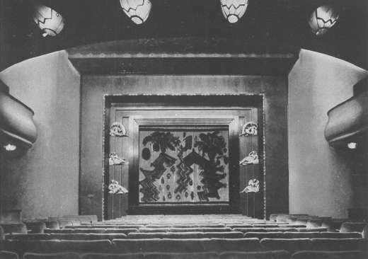 Photograph of a cinema auditorium looking towards the stage: patterned hanging lamps, curved pale side walls, and an angular painted stage curtain above rows of seats