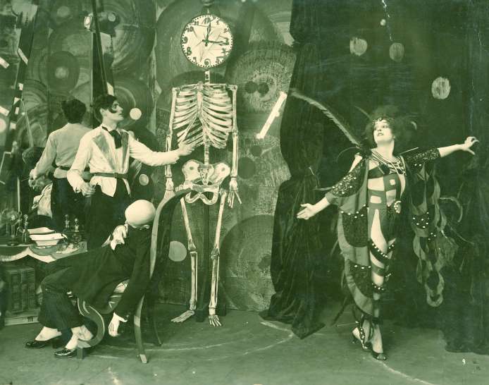 Production still: a man in waistcoat and bow tie gestures towards a full skeleton hanging beside a clock, a bald man crouches at the left, and a woman in a beaded costume stands at the right with her arms outstretched against a painted backdrop