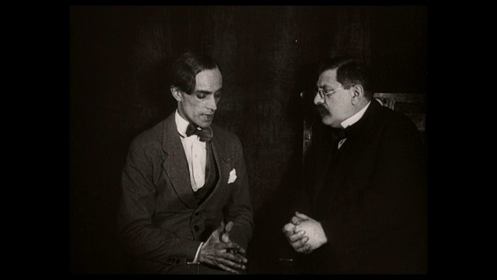 Black-and-white film still, pillarboxed in black. Two men sit close together in a dark panelled room. On the left a gaunt young man in a light checked suit with a bow tie leans forward, head lowered and hands clasped between his knees. On the right an older, heavier man with a walrus moustache and round wire spectacles, in a dark suit and bow tie, turns toward him with his hands folded