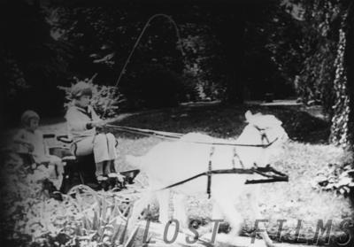 Film still: a child drives a small cart drawn by a white pony along a garden path, a second child seated at the left