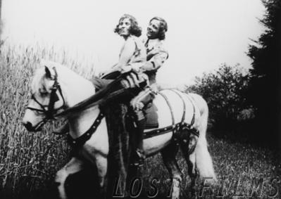 Film still: a young man and a young woman ride together on a white horse through tall grass at the edge of a wood
