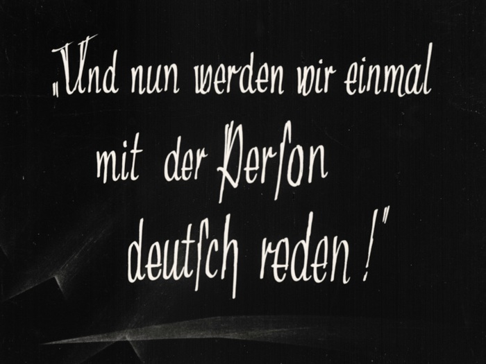 Hand-lettered intertitle from Moral, white script on black, reading "Und nun werden wir einmal mit der Person deutsch reden!"
